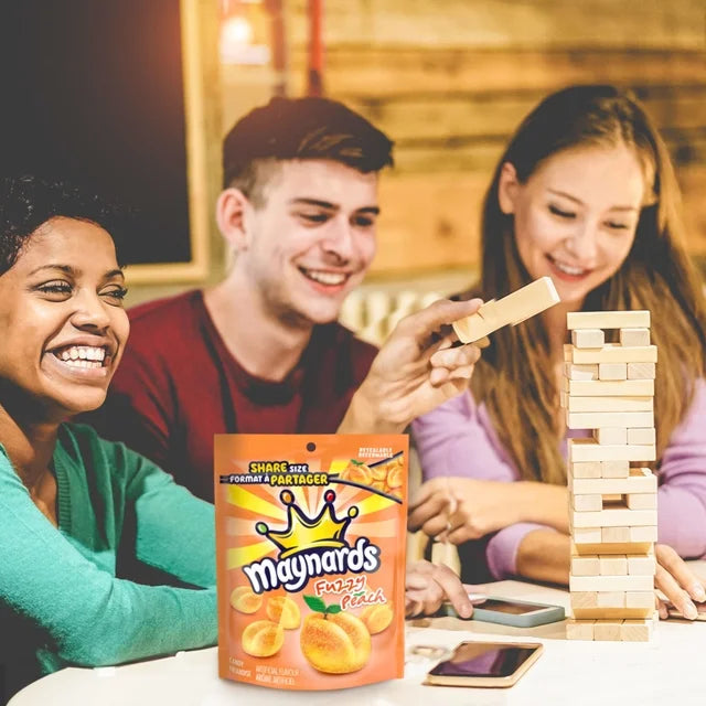 Three people playing with a wooden block tower game, with a Maynards snack packet in the foreground.