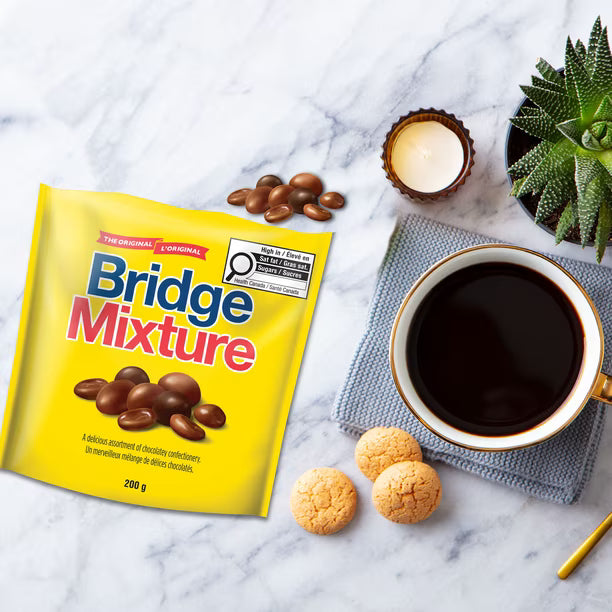 A yellow bag of Bridge Mixture chocolate-coated cookies is styled on a white marble surface next to a cup of black coffee, three cookies, a small candle, and a succulent plant.